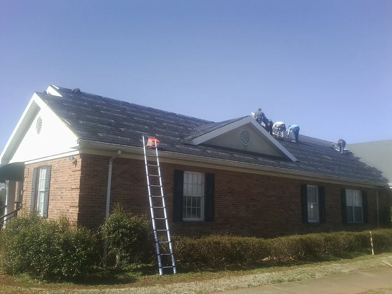 Crew tearing shingles off a brick ranch roof with a ladder set — Emergency Roof Repair work by our crew in Deltona